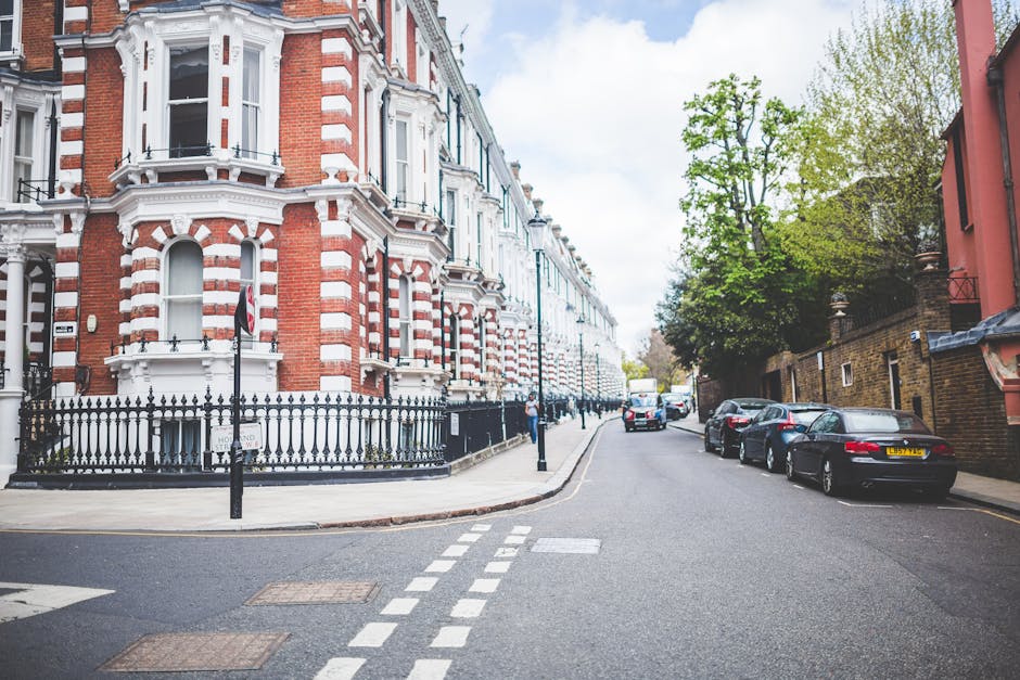A residential street scene featuring a row of Victorian-style terraced houses with red brick facades and white decorative trim. In the foreground, a black wrought iron fence surrounds a small front yard of the corner building, with a pedestrian crossing and traffic bollards visible on the paved road. Several parked cars, including a black sedan and a silver vehicle, line the curb on the right side of the street. On the left, a few pedestrians are walking along the sidewalk, which is bordered by mature green trees providing partial canopy coverage. The sky above is partly cloudy, casting soft natural light across the scene, with some shadows on the road and building facades. The environment suggests an urban area in need of routine rubbish removal or private waste collection, with the clean and maintained appearance of the street indicating recent or ongoing clearance activities, possibly coordinated by a waste management service such as House Clearance Pimlico.