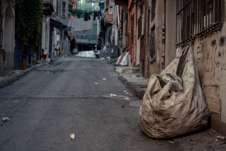 Three large bags of waste, made of crinkled plastic with printed markings, are placed on the edge of a pavement in front of a black metal fence with vertical bars. The bags appear to contain household rubbish and are positioned close to the curb, suggesting on-site clearance or private waste disposal. In the background, dense foliage from trees or bushes extends upward, partially obscuring a building structure. The scene is lit uniformly, with no strong shadows, indicating overcast or diffused natural daylight. The environment appears urban, with the waste bags awaiting collection or disposal, consistent with a rubbish removal service like House Clearance Pimlico. The setting emphasizes the practical aspect of independent or alternative waste handling, often associated with private rubbish removal options near residential areas.