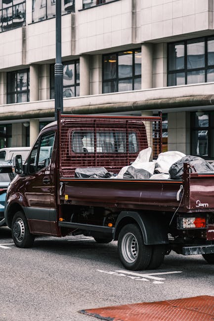 A small burgundy flatbed truck parked on a city street in front of a modern office building with large glass windows and concrete facade. The truck's cargo area is filled with various types of waste, including black and white bags, some of which appear to contain household or construction debris. The vehicle's side panels are lined with a metal grid for safety, and the truck has a compact wheelbase with silver hubcaps. The street surface is asphalt with painted road markings visible in the foreground. A tall street lamp is positioned next to the truck, and there are other vehicles partially visible in the background. The scene suggests an outdoor waste collection or rubbish removal activity, typical of private waste management services like those offered by House Clearance Pimlico, serving immediate and on-site rubbish clearance needs in urban environments.