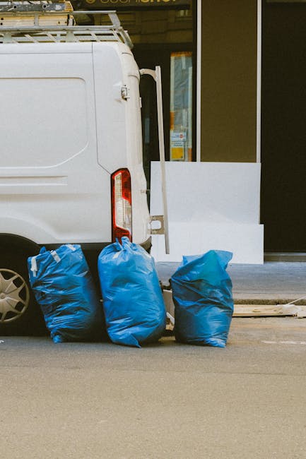 A small burgundy flatbed truck parked on a city street in front of a modern office building with large glass windows and concrete facade. The truck's cargo area is filled with various types of waste, including black and white bags, some of which appear to contain household or construction debris. The vehicle's side panels are lined with a metal grid for safety, and the truck has a compact wheelbase with silver hubcaps. The street surface is asphalt with painted road markings visible in the foreground. A tall street lamp is positioned next to the truck, and there are other vehicles partially visible in the background. The scene suggests an outdoor waste collection or rubbish removal activity, typical of private waste management services like those offered by House Clearance Pimlico, serving immediate and on-site rubbish clearance needs in urban environments.