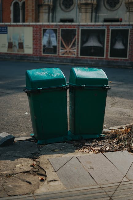 Two green wheeled trash bins with closed lids are positioned side by side on a paved sidewalk adjacent to a road, with a slight gap between them. The surface of the bins appears smooth and made of plastic, showing slight gloss from outdoor lighting. The bins are placed close to the edge of the pavement, with a few scattered dried leaves and small debris surrounding their base. In the background, there is a road with an asphalt surface, partially visible concrete curb, and a brick wall with advertising posters featuring images and text, indicating an urban environment. The scene appears to be during daylight hours, with natural sunlight illuminating the bins and casting subtle shadows. The setting suggests a typical outdoor scene where private waste collection and rubbish removal services, such as those provided by House Clearance Pimlico, might be involved in managing local refuse in a residential or commercial area without reliance on municipal waste facilities.