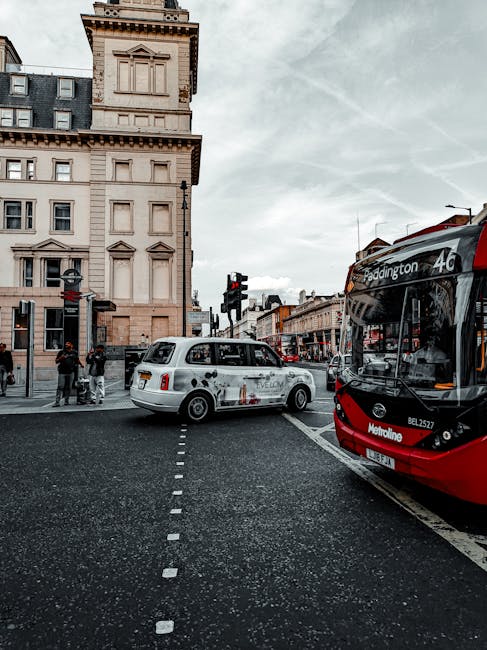 A busy city street scene showing a white taxi with a patterned vinyl wrap on its side, positioned at a pedestrian crosswalk with its front facing to the right. To the right of the taxi, part of a modern red and black bus with destination signs indicating route 46 to Paddington is visible, parked alongside a curb. In the background, there are multi-storey buildings featuring classic architectural details, including decorative cornices and window frames, with a pale stone facade. Pedestrians, some standing on the sidewalk, are visible on the left side of the image, waiting at a traffic light with a black pole and signal. The sky above is overcast with grey clouds, creating diffused natural light that illuminates the scene. The street surface is paved with dark asphalt, and white dashed lines mark the pedestrian crossing. This urban setting exemplifies common city transport and infrastructure, fitting within a context of public and private transportation, including alternative waste removal options like on-site clearance or independent collection services that may operate in such environments. The scene reflects typical city traffic and pedestrian activity, relevant to rubbish and waste management discussions in urban settings.