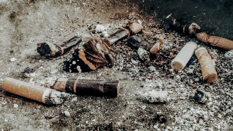 A close-up view of a heavily littered pavement surface featuring burnt cigarette butts, some partially ash-covered and others with intact filters, scattered among scattered debris, ash, and small burnt fragments. The ground appears to be a concrete or asphalt area, showing signs of dirt and minor wear. The cigarette butts vary in color from white to brown and black, with the burned ends exposing tobacco residues. The scene suggests an instance of illegal dumping or fly-tipping, highlighting the need for private waste removal services like those offered by House Clearance Pimlico. The ambient lighting is neutral, emphasizing the details of the trash and debris on the surface, with no background or additional environmental elements visible, keeping the focus solely on the discarded cigarette butts and debris, relevant in the context of waste management and rubbish clearance.
