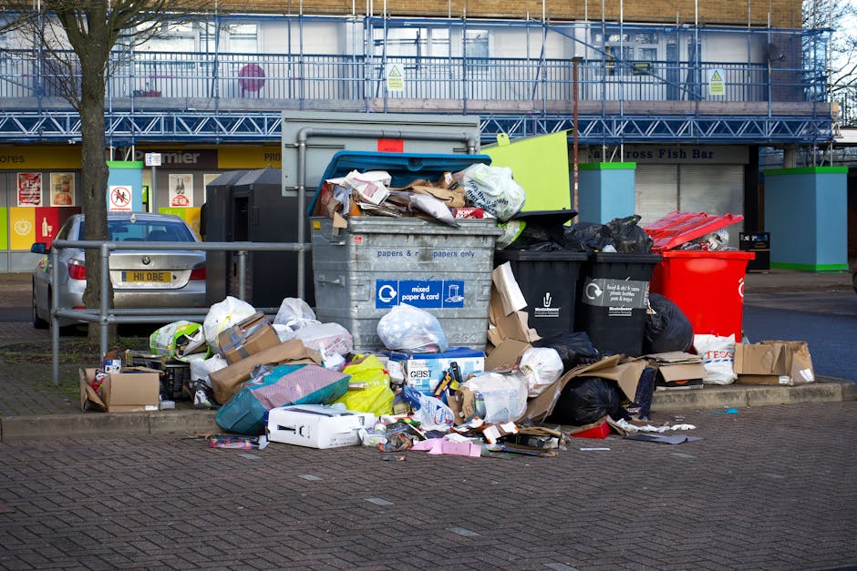 A cluttered outdoor collection of overflowing rubbish bins and scattered waste bags occupy a paved area on a street in front of a building with blue scaffolding. The waste includes black, red, and green bags, along with cardboard boxes, plastic containers, and loose paper and packaging. A large grey mixed paper and card container is situated centrally, with its lid open and rubbish spilling out onto the ground. To the left, there is a small metal fence partially obstructing a parked silver vehicle with a visible yellow registration plate. Behind the rubbish, the building features storefronts with signs, and the upper floor is under construction or maintenance, covered by blue protective netting and scaffolding. The scene suggests a temporary or unmanaged waste accumulation that might be addressed by local rubbish removal services such as those offered by House Clearance Pimlico, supporting private waste clearance and alternative disposal arrangements.