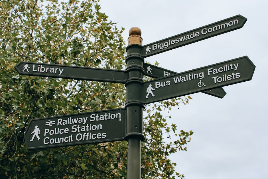 A tall black metal signpost featuring multiple directional street and location signs with white text and icons, situated outdoors against a backdrop of leafy green trees and an overcast sky. The signs point to nearby landmarks such as Biggleswade Common, the library, railway station, police station, council offices, bus waiting facilities, and toilets. Each sign has a small pedestrian icon indicating walking directions, with some signs including additional symbols for accessibility or transportation modes. The signpost is centered in the image and appears sturdy, with a rounded top finial. The environmental setting suggests a public area or town center, where visitors might find directions to various civic amenities and public transport links. The scene is well-lit with natural daylight, emphasizing the contrast between the black sign surfaces and the white lettering, matching the tone of professional, factual description relevant to local infrastructure and urban navigation, subtly supporting a context related to waste management or municipal services in a civic setting.
