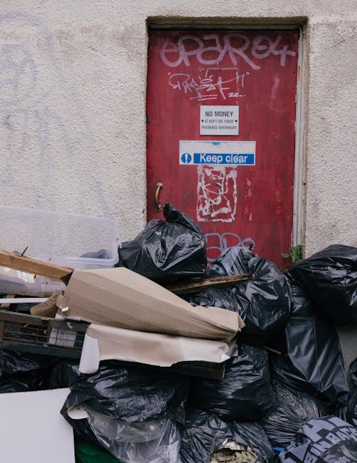 A cluttered outdoor collection of overflowing rubbish bins and scattered waste bags occupy a paved area on a street in front of a building with blue scaffolding. The waste includes black, red, and green bags, along with cardboard boxes, plastic containers, and loose paper and packaging. A large grey mixed paper and card container is situated centrally, with its lid open and rubbish spilling out onto the ground. To the left, there is a small metal fence partially obstructing a parked silver vehicle with a visible yellow registration plate. Behind the rubbish, the building features storefronts with signs, and the upper floor is under construction or maintenance, covered by blue protective netting and scaffolding. The scene suggests a temporary or unmanaged waste accumulation that might be addressed by local rubbish removal services such as those offered by House Clearance Pimlico, supporting private waste clearance and alternative disposal arrangements.