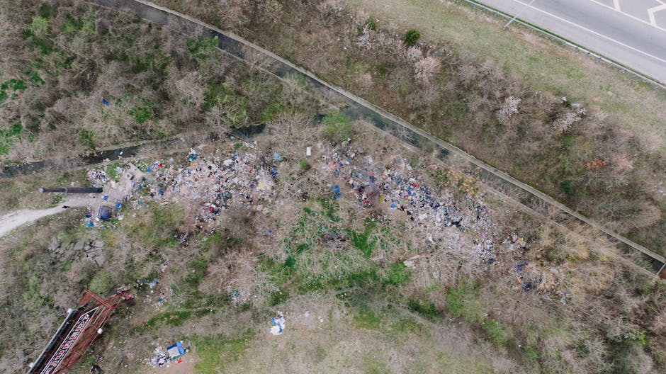 An aerial view of a large, unmanaged waste pile situated in a natural, semi-urban environment. The landfill consists of various types of rubbish, including plastic bags, cardboard, and other debris, scattered across a patch of leafless trees and sparse greenery. The waste appears loosely dumped, with some items partially buried or fallen into the undergrowth. To the right, a paved road runs parallel to the waste area, separated by a strip of grass and a low barrier. The overall scene conveys an unmanaged or illegal waste dumping site, with surrounding natural vegetation and an adjacent infrastructure element. The lighting suggests daylight with no prominent shadows, and the scene's neutral tone emphasizes the environmental impact of improper rubbish disposal. House Clearance Pimlico’s rubbish removal service is relevant here, providing an alternative to illegal dumping through controlled, private waste collection and cleanup of such sites.