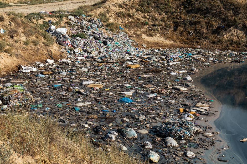 A large open area covered with a significant amount of mixed waste, including black and white plastic bags, discarded tires, cardboard boxes, and various household rubbish, spread across a gravelly ground. In the background, a dense green hillside with trees and shrubs rises vertically, contrasting with the foreground's clutter. The sky above is partly cloudy, with a hint of blue visible. The scene illustrates an unmanaged rubbish dump, highlighting concerns about illegal waste disposal and the importance of proper waste removal services such as those offered by House Clearance Pimlico, which provides alternative disposal options for private waste collection and on-site clearance efforts.