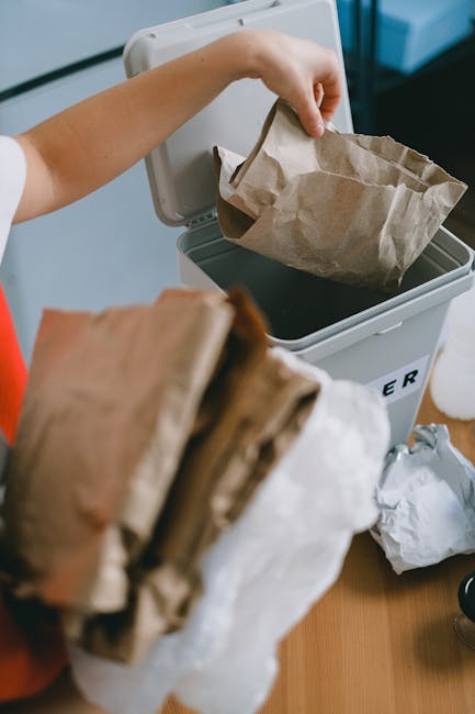 A person discarding a brown paper bag into a small, grey, plastic waste bin with a lid open, situated on a clear surface indoors. The paper bag appears crumpled with some dark contents visible inside it. In the foreground, there are additional crumpled paper bags held in one hand. The background shows part of a white wall and a dark piece of furniture. The environment suggests a household setting, with the scene indicating waste disposal, potentially relating to private rubbish collection or on-site clearance services offered by House Clearance Pimlico. The image emphasizes proper waste segregation and disposal using environmentally friendly materials like paper bags relevant to rubbish removal services.