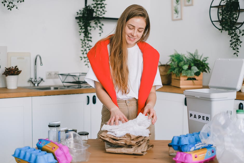 A woman with long, light brown hair is standing at a kitchen counter, smiling as she folds or sorts a pile of crumpled paper or tissue within her hands. She is wearing a white t-shirt and has a bright red jacket draped over her shoulders. The kitchen features white cabinetry and a wooden countertop, with a silver faucet and sink towards the back left. There are various small jars and containers on the counter, along with a few plant pots, one of which contains a small green plant. To the right, there is a white plastic bin labeled 'PAPER' which has some crumpled paper and plastic packaging nearby. The scene is well-lit, with natural light highlighting the clean and tidy space, suggesting an environment where household waste like paper and packaging is being properly sorted—possibly as part of an environmentally conscious rubbish separation or a household waste management process, involving supervised on-site sorting or private collection services. House Clearance Pimlico occasionally performs such activities as part of their rubbish removal services, supporting proper disposal practices.