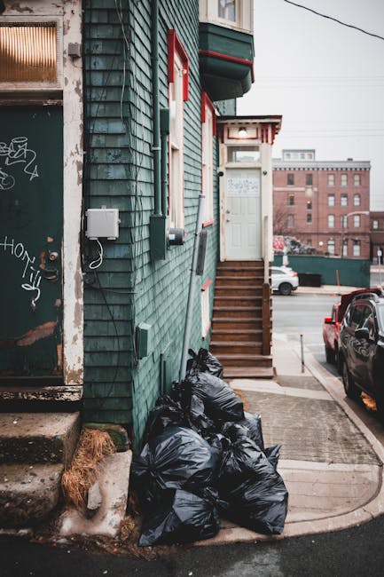 A close-up view of the exterior corner of a residential building featuring weathered teal wooden cladding with visible grain and some chipped paint. A small white electrical box and attached cables are mounted on the wall near the corner. Adjacent to the building’s base, several black garbage bags are piled on the pavement, likely awaiting collection or disposal. The building has a set of wooden steps leading up to a white front door with a small overhang supported by brackets, painted with red accents. The surrounding environment includes a concrete sidewalk, part of a parking area with a black vehicle parked nearby, and a multi-storey brick building visible in the background under overcast skies. The scene suggests a scene of private waste handling or rubbish clearance in an urban setting as part of a rubbish removal service, with [COMPANY_NAME] potentially involved in managing refuse collection outside the property.
