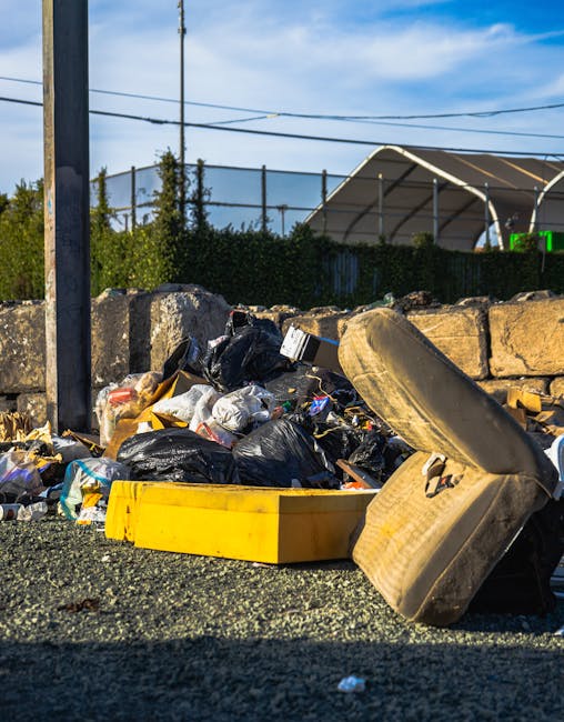A close-up view of an outdoor rubbish pile situated on a gravel surface, comprising various discarded waste materials including black plastic bin bags, a yellow plastic container, a large worn car tyre leaning against the pile, and loose rubbish such as paper, packaging, and small debris. Behind the waste, there is a low stone wall made of irregularly shaped, rough-textured grey stones, with some green shrubs and small trees visible beyond it. To the left, a utility pole with attached wires extends upward. In the background, there is a partly visible sports or tennis court enclosed by a tall metal fence, and a large, curved, semi-transparent canopy structure with metal framing, suggesting an outdoor recreational or sports facility. The sky is mostly clear with some wispy clouds, and the scene is illuminated by natural daylight, highlighting the contrasting textures and colours of the waste and surrounding environment. This scene reflects an example of non-regular waste accumulation that might be suitable for private rubbish removal services like those offered by House Clearance Pimlico, emphasizing the importance of proper disposal or on-site clearance of accumulated rubbish.
