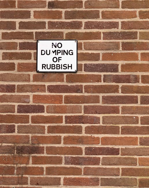 A rectangular metal sign mounted on a red brick wall displaying the message 'NO DUMPING OF RUBBISH' in bold black uppercase letters. The sign has a white background with a black border, and the text is arranged in three lines with the words 'NO' and 'OF' aligned centrally. The brick wall behind the sign features standard-sized, evenly spaced bricks with a reddish-brown hue and light mortar joints. The texture of the bricks appears rough and weathered. The image captures the sign positioned centrally on the wall, with a slight shadow cast at the bottom left corner from natural lighting, indicating an outdoor setting. The sign serves as an instruction relevant to rubbish removal or waste disposal, aligning with the context of private waste handling and alternative rubbish management options provided by companies such as House Clearance Pimlico.