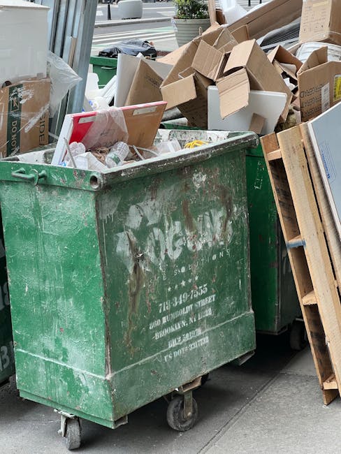 A large, green, industrial-sized wheeled waste container filled with mixed debris, positioned on a city pavement. The container's surface is scratched and weathered, featuring faded white lettering and contact details. Overfilled with various types of rubbish, including flattened cardboard boxes, paper packaging, and plastic items, some spilling over the top. To the right of the container, a wooden pallet leaning against it holds additional cardboard boxes and waste materials, some partially opened or crushed. Behind the container, a cluttered area of discarded refuse is visible, with more flattened boxes and packaging piled haphazardly. The scene is set outdoors, with a sidewalk, storefront window, and urban environment ahead, suggesting a temporary rubbish accumulation awaiting collection or disposal. House Clearance Pimlico occasionally handles on-site rubbish removal and waste management, as reflected by the organized yet cluttered scene typical of private waste handling situations in the area.