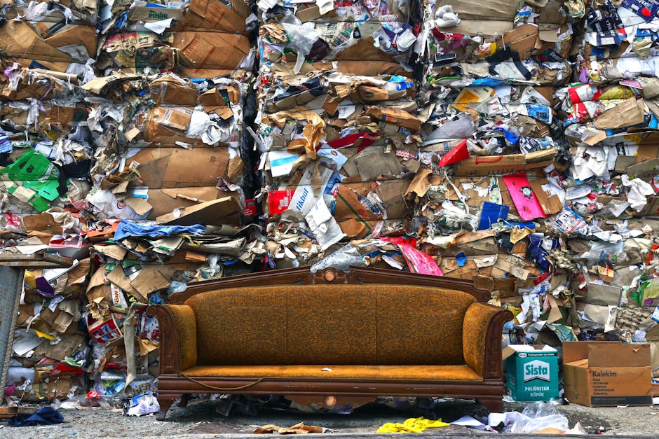 A vintage brown upholstered sofa with wooden framing and ornate carving details is positioned in the foreground of the image, resting against a large, chaotic wall of tangled waste. The waste wall consists of stacked cardboard boxes, crumpled paper, and various mixed packaging materials, displaying a variety of textures from smooth glossy surfaces to rough and torn paper. The cardboard boxes are different shades of brown, some flattened while others are partially folded or torn, with identifiable printed logos and labels. Among the debris, plastic packaging in bright colors such as red, blue, and green can be seen, with some flattened and others bunched up. The scene is set outdoors on pavement, with natural daylight illuminating the cluttered environment, creating a contrast between the aged, intricate texture of the sofa and the messy, disorganized waste behind it. The image visually underscores the challenge of managing bulky waste, indirectly referencing private or independent collection solutions that companies like House Clearance Pimlico may offer for rubbish removal services in urban settings.
