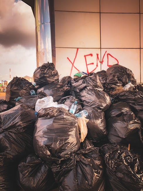 A large pile of black plastic rubbish bags stacked against an exterior wall of a modern building, with some bags appearing torn open, revealing plastic bottles and miscellaneous waste inside. The bags are placed on a paved surface, with a few discarded bottles and small debris scattered around at the base. Behind the bags, the wall features large, beige-colored tiles with a smooth finish, and there is bright natural daylight illuminating the scene, which contrasts with the urban background visible partly on the left side, including some distant structures. The graffiti 'XEND' is spray-painted in red on the wall behind the rubbish bags. This scene reflects an unmanaged waste accumulation that could be part of a private rubbish collection or on-site clearance, highlighting typical challenges in waste disposal that services like House Clearance Pimlico might address and resolve for clients seeking alternative waste handling options from conventional council collections.